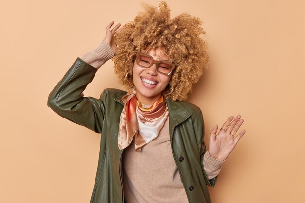 Une femme heureuse et détendue avec des cheveux blonds bouclés et touffus se dresse sur un fond beige danse joyeusement à la musique lève les bras vêtus de vêtements à la mode sourit largement se sent libre et insouciante profite de la vie