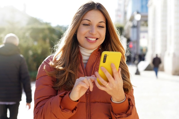 Femme heureuse en doudoune à l'aide d'un téléphone intelligent jaune dans la rue de la ville