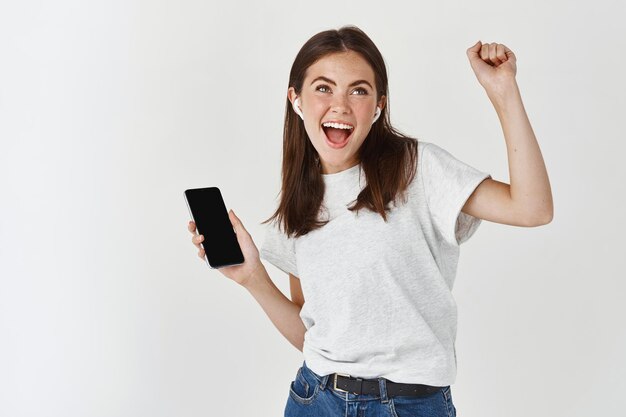 Femme heureuse écoutant de la musique dans des écouteurs sans fil et tenant un smartphone, dansant et chantant sur un mur blanc