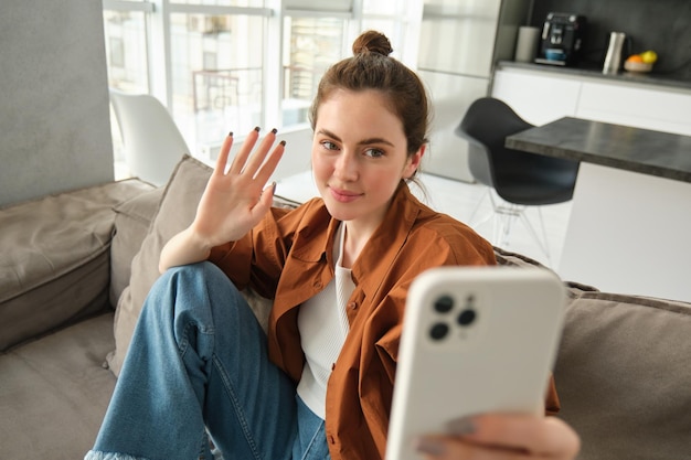 Une femme heureuse est assise à la maison avec un smartphone en ligne en discutant en agitant la main sur le téléphone portable en disant