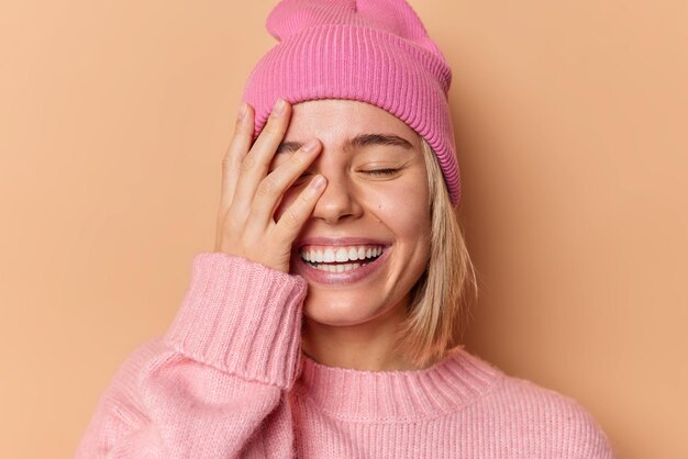 Une femme heureuse fait des sourires de paume au visage montre largement les dents blanches ferme les yeux de satisfaction se sent très heureuse d'entendre d'excellentes nouvelles porte un chapeau et un pull isolés sur fond marron. Notion d'émotions