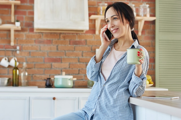 Femme heureuse habillée avec désinvolture, discute avec un ami sur téléphone intelligent, boit des boissons dans la cuisine