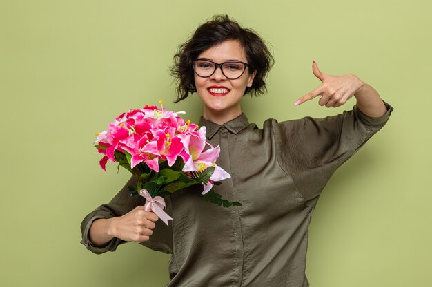 Femme heureuse et heureuse aux cheveux courts tenant un bouquet de fleurs pointant avec l'index vers elle souriant joyeusement célébrant la journée internationale de la femme le 8 mars debout sur fond vert