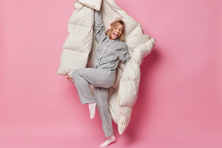 Photo gratuit une femme heureuse et heureuse danse et s'amuse à écouter de la musique via des écouteurs sans fil tient des idiots de couverture autour se sent énergique après un sommeil sain isolé sur fond rose. notion de réveil