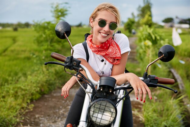 Une femme heureuse et heureuse est assise sur sa moto, heureuse de gagner la compétition de motards, satisfaite des bons résultats, aime la vitesse et le mouvement en plein air. Gens, mode de vie actif et activités de plein air