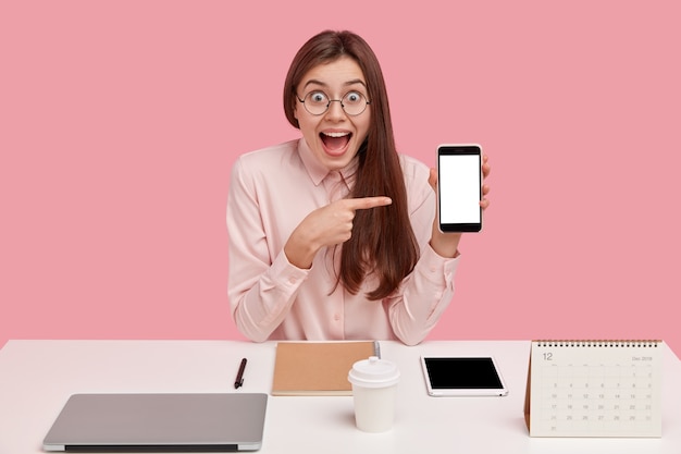 Une femme heureuse et heureuse montre un téléphone portable avec une maquette d'écran, a étonné l'expression du visage, est perfectionniste, s'efforce d'atteindre la perfection