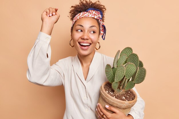 Une femme heureuse et insouciante danse avec le bras levé tient un cactus dans un pot prend soin des plantes d'intérieur sourit largement isolée sur un mur beige