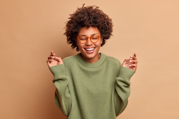 Une femme heureuse lève les mains a une expression joyeuse insouciante ferme les yeux sourit à pleines dents porte des lunettes optiques et un pull isolé sur un mur beige