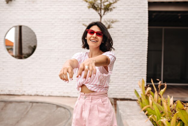 Femme Heureuse à Lunettes De Soleil Posant Sur Rue Avec Les Mains Tendues. Plan Extérieur D'une Femme Bronzée En T-shirt Rose.