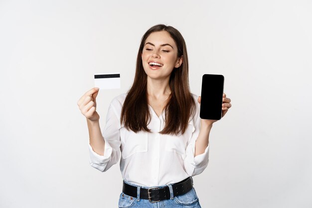 Femme heureuse montrant la carte de crédit et l'écran du smartphone, concept d'achat en ligne, achat dans l'application, debout sur fond blanc.