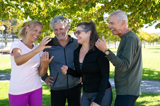 Femme heureuse montrant l'écran du téléphone portable aux camarades du club de remise en forme. Joyeux amis matures debout ensemble après les exercices du matin dans le parc. Concept de retraite ou de communication