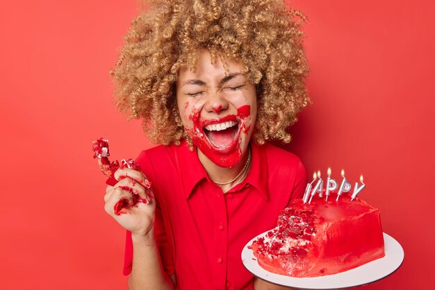 Une femme heureuse optimiste rit avec joie garde la bouche largement ouverte aime manger un délicieux gâteau reçu comme cadeau le jour de la Saint-Valentin enduit de crème isolé sur fond rouge aime la célébration