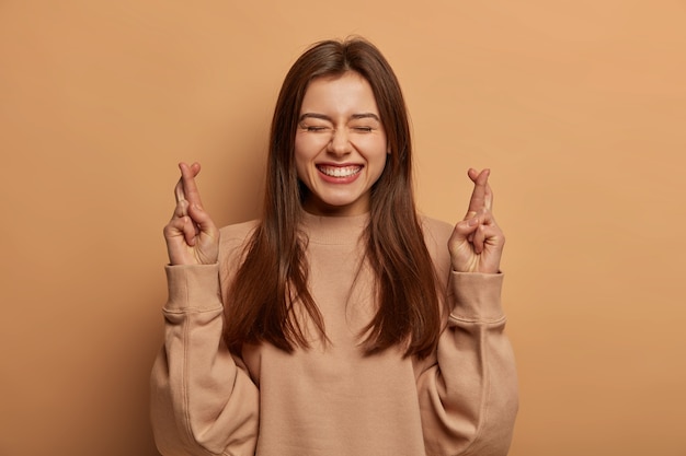 Une femme heureuse et paisible croise les doigts pour avoir de la chance, croit en la bonne chance, anticipe la réalisation des rêves, sourit largement, porte un sweat-shirt marron