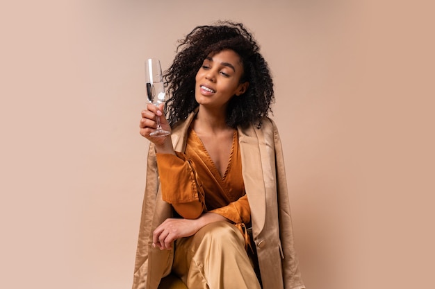 Femme heureuse à la peau bronzée avec des cheveux bouclés parfaits tenant un verre de vin, portant un chemisier orange ielegant et un pantalon en soie assis sur un mur beige chaise vintage.