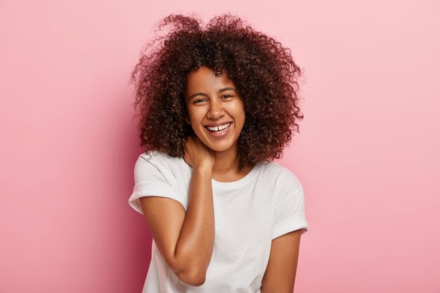 Une femme heureuse à la peau sombre a une conversation amusante, regarde avec un large sourire, divertit par un ami, a une expression de visage ravie, habillée de vêtements de tous les jours, se tient à l'intérieur. Concept d'émotions