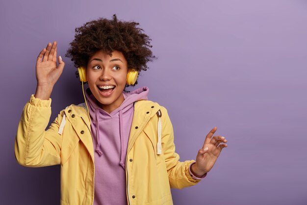 Une femme heureuse à la peau sombre danse tout en entendant une musique agréable dans les écouteurs, regarde quelque part pensivement, écarte les bras, se sent heureuse