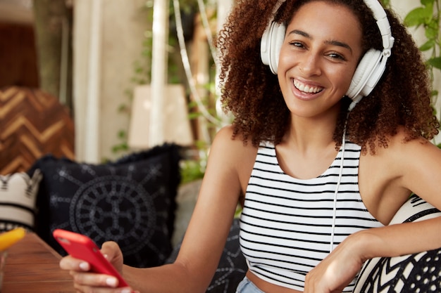 Une femme heureuse à la peau sombre écoute la chanson préférée dans les écouteurs, discute en ligne sur un téléphone intelligent, porte un t-shirt à rayures décontracté, télécharge des pistes populaires dans une liste de lecture. Femme africaine divertit au café