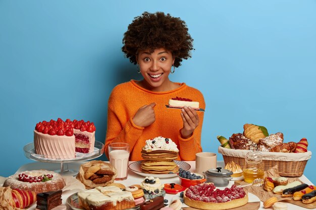 Une femme heureuse à la peau sombre a un regard positif, se montre du doigt, tient un délicieux morceau de gâteau, demande si elle doit tout manger, vêtue d'un pull orange, isolée sur un mur bleu.
