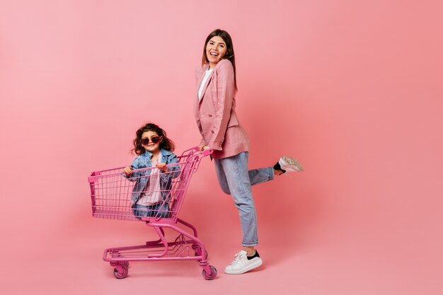 Femme heureuse avec petite fille posant après le shopping. Mère souriante debout près du chariot du magasin.