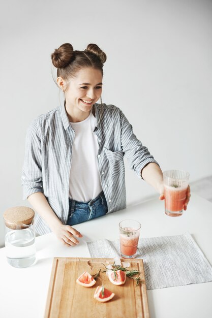 Femme heureuse avec des petits pains souriant étirement du verre avec du smoothie détox de pamplemousse à quelqu'un. Fond de mur blanc. Aliments diététiques sains.