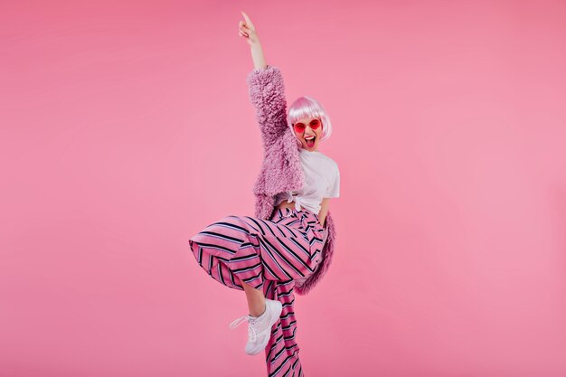 Femme heureuse porte un pantalon rayé et une perruque rose riant pendant la séance photo. Confiant jeune femme en lunettes de soleil et veste moelleuse danse drôle