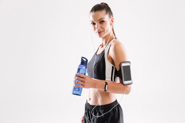 Femme heureuse de remise en forme dans les écouteurs debout et tenant une bouteille d'eau