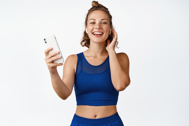 Femme heureuse de remise en forme dans les écouteurs, tenant le smartphone et riant. Entraînement avec musique sur téléphone portable, portant des vêtements de sport, fond blanc.