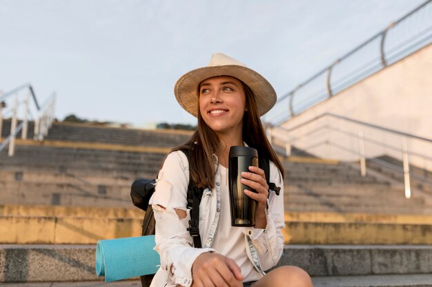 Femme heureuse avec sac à dos et chapeau tenant thermos lors d'un voyage