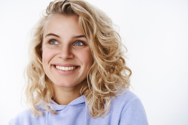 Photo gratuite femme heureuse se rappelant de beaux souvenirs chaleureux et tendres souriante ravie en regardant avec de beaux yeux bleus dans le coin supérieur gauche comme souvenir d'un beau moment sur un mur blanc