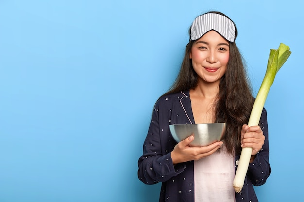 Photo gratuite femme heureuse se réveille tôt le matin pour préparer le petit-déjeuner, vêtue de vêtements de nuit confortables, détient un bol en acier et poireau vert