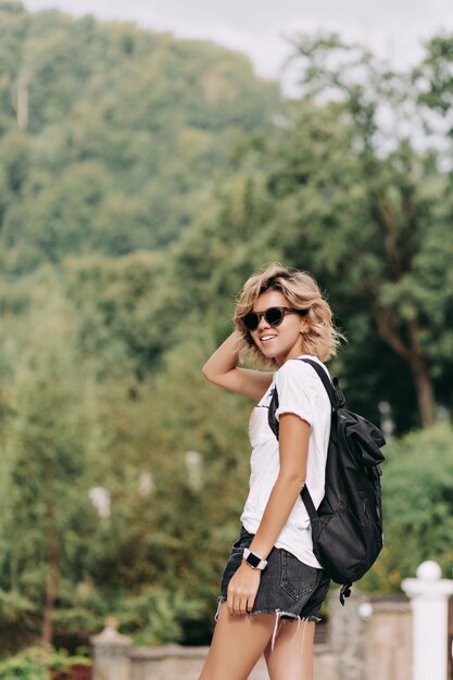 Femme heureuse souriante avec une coiffure courte habillée t-shirt blanc et un short en lunettes de soleil noires voyageant dans les montagnes, bonne journée ensoleillée, randonnée dans les montagnes