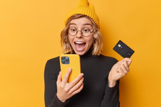 Une femme heureuse surprise porte un chapeau et un col roulé noir décontracté regarde l'écran de l'application smartphone détient la carte de crédit utilise un compte bancaire pour payer en ligne isolé sur fond jaune obtient une remise en argent