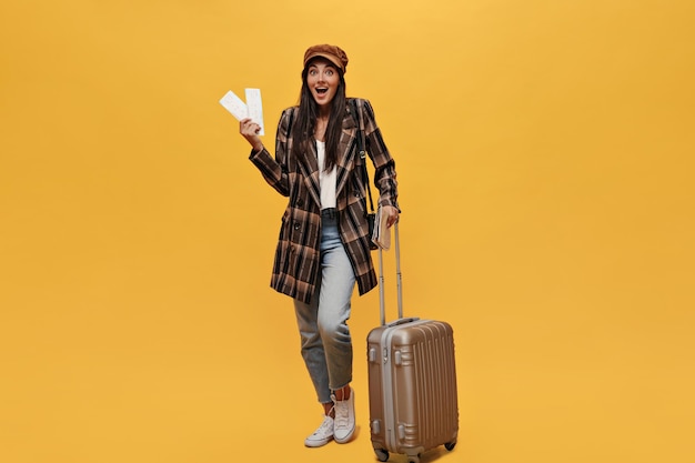 Une femme heureuse surprise regarde dans la caméra et détient des billets Fille bronzée aux cheveux longs en manteau et jeans pose avec un sac de costume sur fond jaune