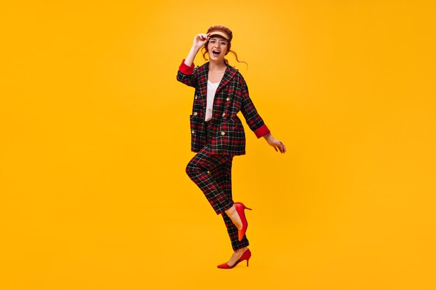 Une femme heureuse en tenue à carreaux se déplace sur fond orange Une fille joyeuse en casquette marron et lunettes a un ventilateur sur fond isolé