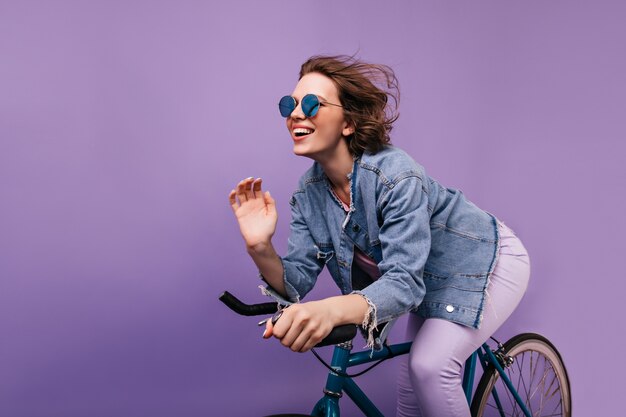 Femme heureuse en veste décontractée posant sur le vélo. Fille aux cheveux courts émotionnelle dans des lunettes scintillantes à vélo.
