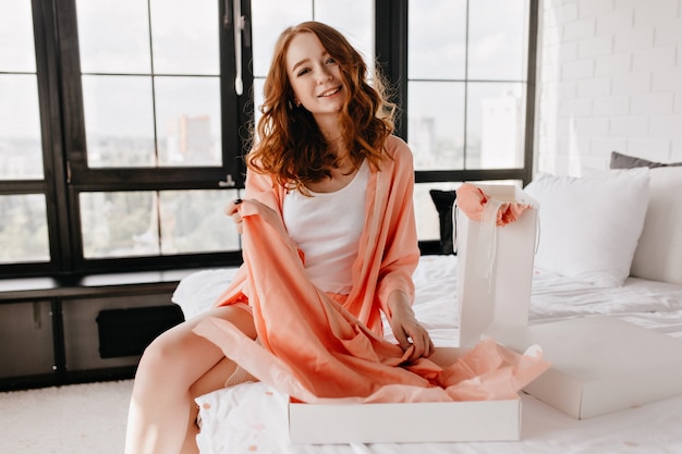 Femme heureuse en vêtements de nuit souriant dans la chambre. Fille bouclée intéressée assis sur le lit avec une expression de visage heureux.