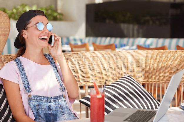 Une femme hipster élégante et joyeuse porte des lunettes de soleil à la mode, une casquette et une salopette en jean, a une conversation agréable via un téléphone portable, est assise sur un canapé confortable contre l'intérieur du café, boit un cocktail