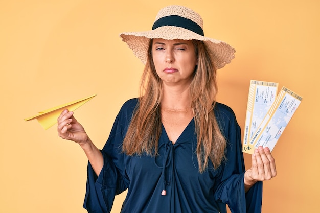 Femme hispanique d'âge moyen portant un chapeau d'été tenant une carte d'embarquement et un avion en papier sceptique et nerveux fronçant les sourcils contrarié à cause d'un problème de personne négative