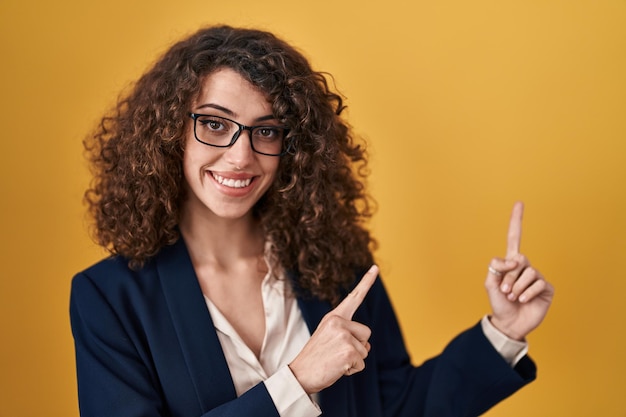 Photo gratuite femme hispanique aux cheveux bouclés debout sur fond jaune souriant et regardant la caméra pointant avec deux mains et doigts sur le côté