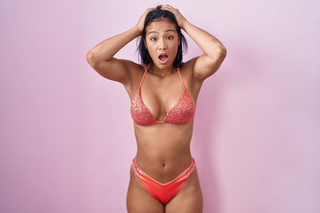 Femme hispanique portant un bikini fou et effrayé avec les mains sur la tête, effrayé et surpris du choc avec la bouche ouverte