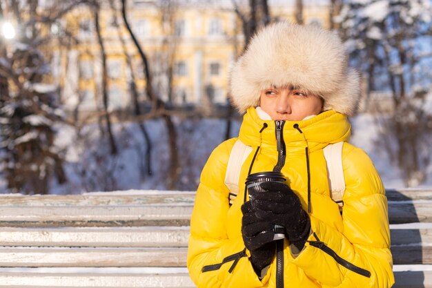 Photo gratuite une femme en hiver dans des vêtements chauds dans un parc couvert de neige par une journée ensoleillée est assise sur un banc et gèle du froid, est malheureuse en hiver, tient le café seul