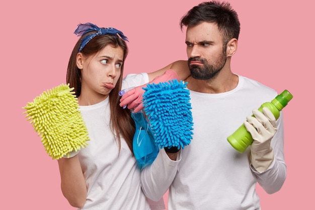 La femme et l'homme européens bouleversés froncent les sourcils tout en se regardant, maintenez le spray et la bouteille de détergent, chiffons colorés