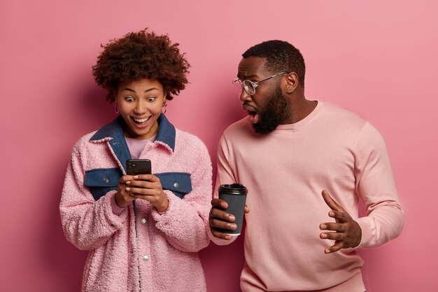 Une femme et un homme noirs émotifs regardent un smartphone, réagissent à de superbes nouvelles, reçoivent un message, boivent du café à emporter