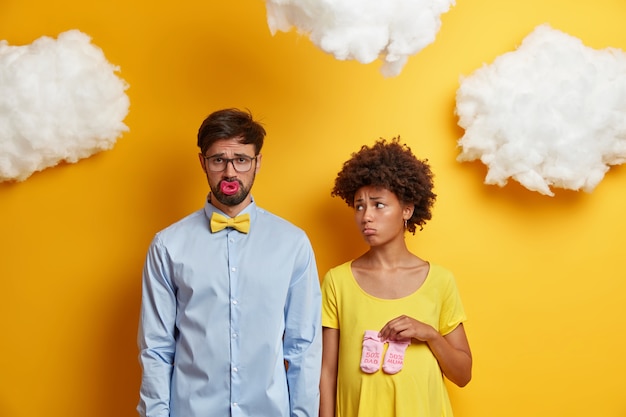 Photo gratuite une femme et un homme tristes attendent bébé, découvrent de mauvaises nouvelles, posent avec un téton et des chaussettes pour enfants, se préparent à devenir parents, se tiennent côte à côte contre le jaune. jeune famille se prépare à la naissance