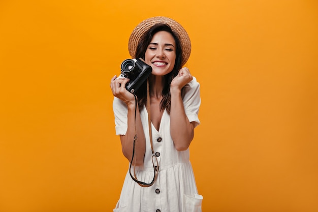 Femme d'humeur joyeuse tenant un appareil photo rétro sur fond orange. Fille merveilleuse dans des vêtements élégants légers et posant un chapeau de paille.