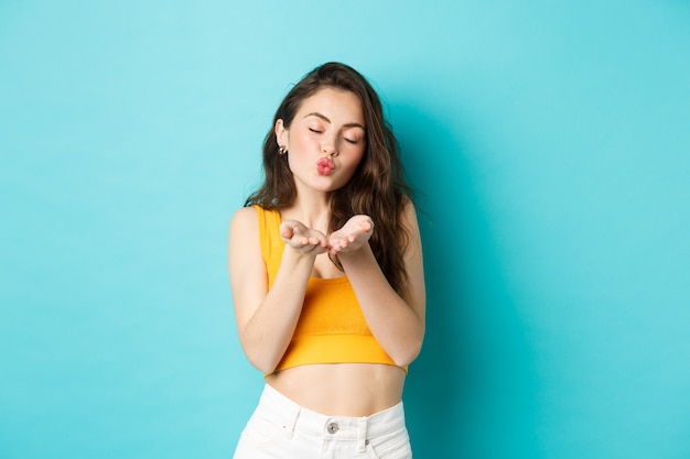 Une femme idiote romantique ferme les yeux et les lèvres plissées, vous souffle un baiser d'air, debout rêveuse sur fond bleu