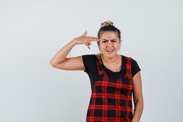 Femme imitant le geste de suicide en robe chasuble et à la vue de face, mélancolique.