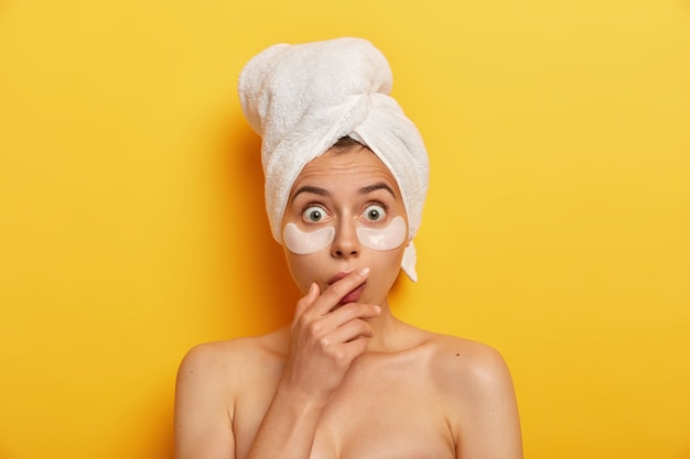 Une femme impressionnante a une beauté naturelle, regarde avec des yeux écarquillés, applique des patchs d'hydrogel qui font une différence visible sur sa peau, une production excessive d'huile, touche la bouche avec les mains