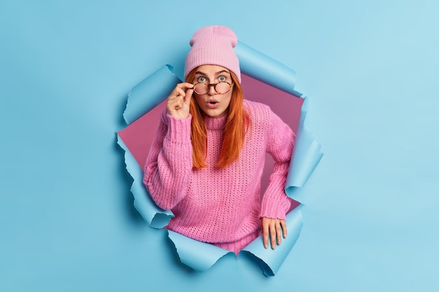 Une femme impressionnée a l'air étonnamment et se sent étonnée, porte un chandail tricoté avec un chapeau rose, perce le papier