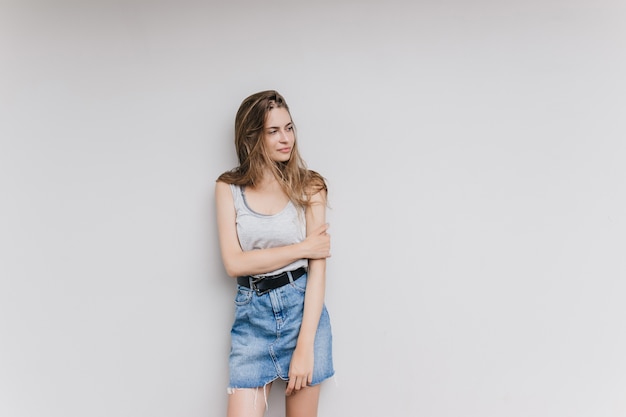 Femme incroyable en jupe en jean posant dans un mur blanc. Tir intérieur d'une jolie fille brune isolée avec une expression de visage pensif.